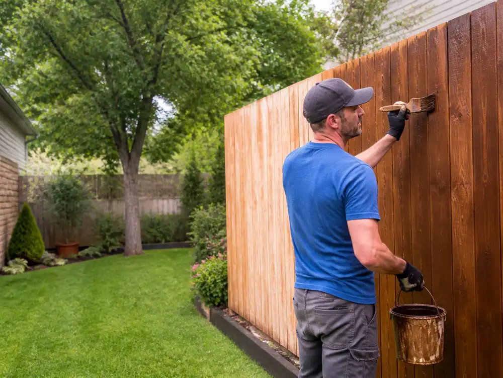 A man in a blue shirt is painting a wooden fence in a backyard.